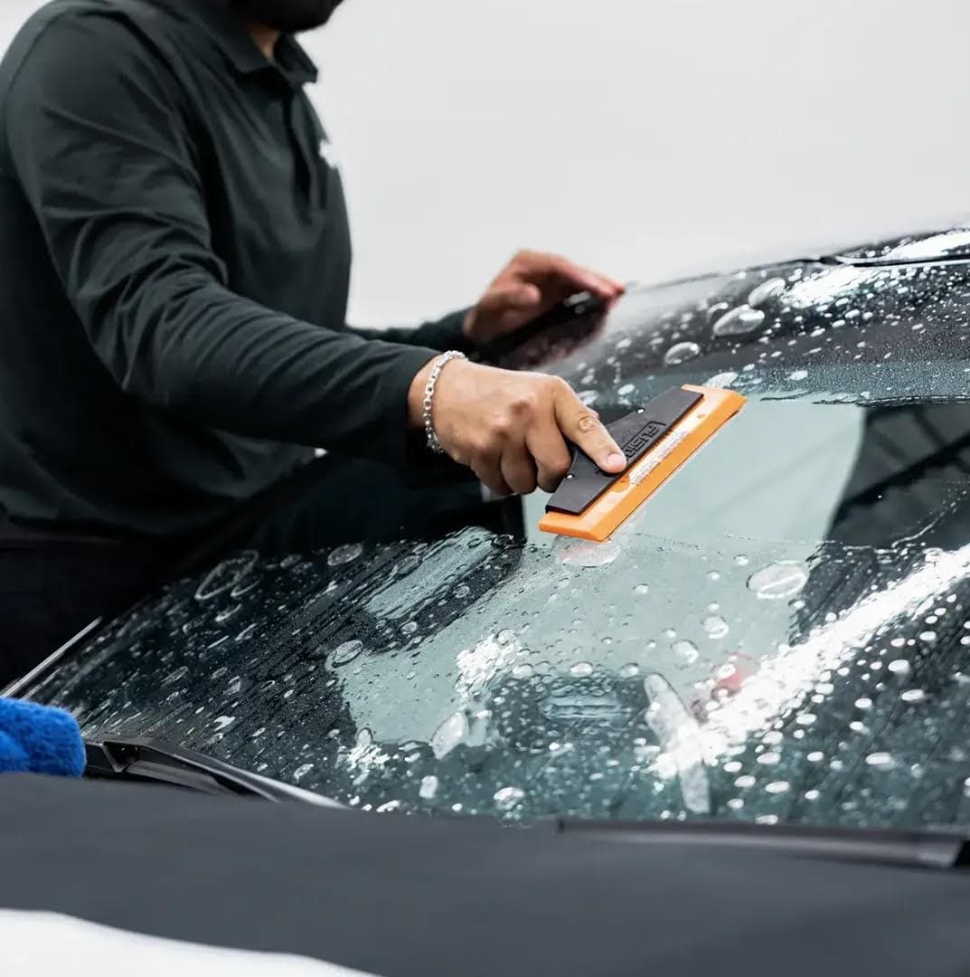 Technician using a squeegee to install clear protective film across a wet vehicle windshield